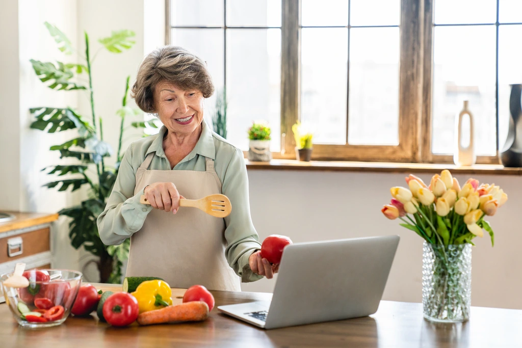 Carla Peterson holding a red apple and a spatula while demonstrating a recipe on a video call in her kitchen.