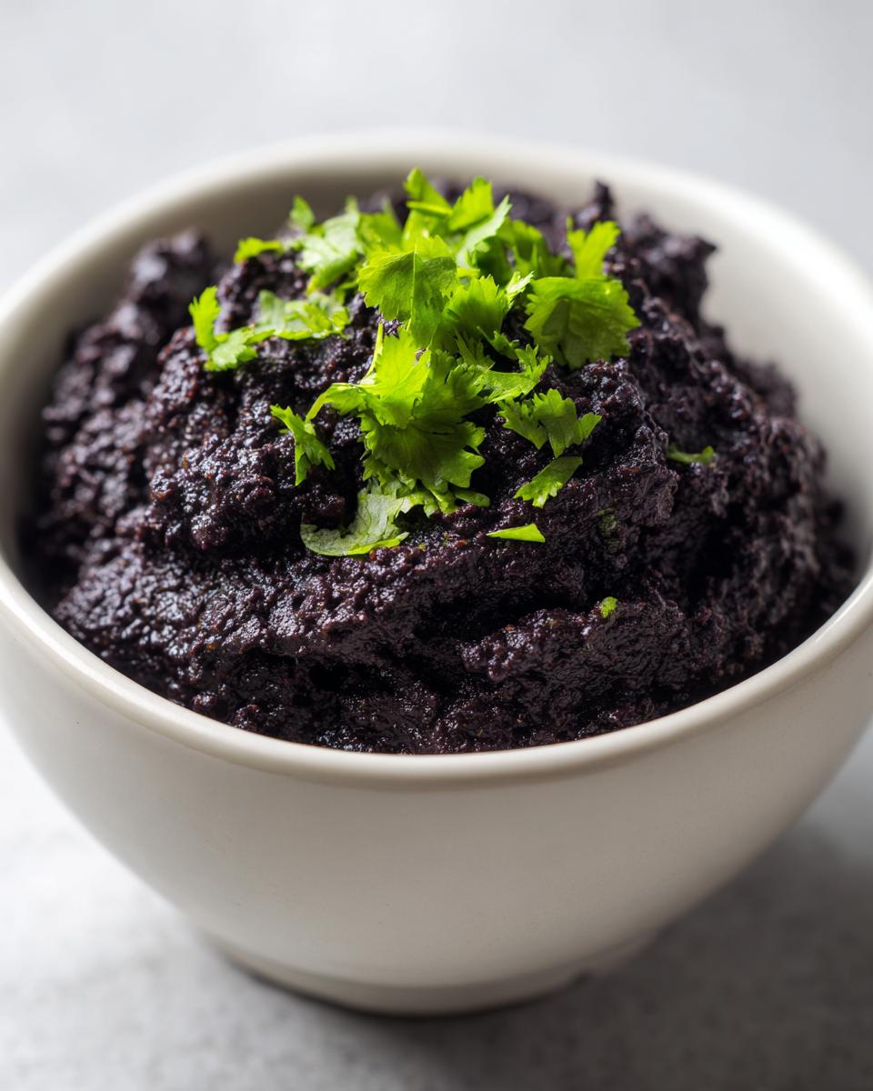 A close-up of thick, dark purple black bean dip served in a white bowl and garnished with fresh cilantro.