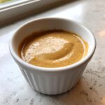 A close-up of thick, creamy, golden-brown peanut sauce served in a small white fluted ramekin.