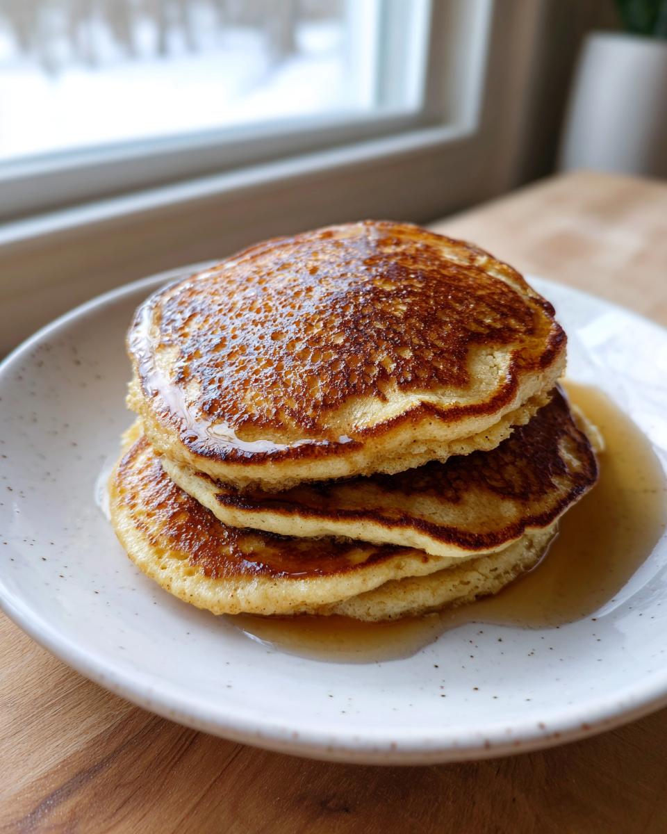 A close-up stack of three golden brown almond flour pancakes drizzled with syrup on a speckled white plate.