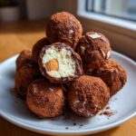 A pile of rich almond joy truffles dusted heavily with cocoa powder, one is cut open showing the coconut filling and whole almond.