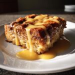 A close-up of a rich, baked bread pudding slice topped generously with a warm caramel sauce.