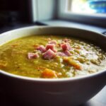 Close-up of a thick, green pea soup in a white bowl, garnished with diced ham and visible pieces of carrot.