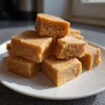 A stack of creamy, light brown squares from a peanut butter fudge recipe sprinkled with fine sugar crystals.