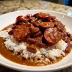 Close-up of a white bowl filled with white rice topped generously with rich, savory red beans and rice and thick slices of andouille sausage.