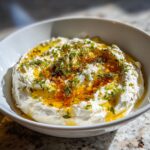 Close-up of a creamy white ricotta dip generously drizzled with olive oil and sprinkled with fresh green herbs.