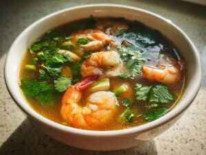 A white bowl filled with rich broth, large pink shrimp, and fresh green herbs, showcasing the amazing tom yum soup.