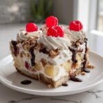A delicious slice of banana split cake topped with whipped cream, maraschino cherries, nuts, and chocolate drizzle.