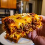 Close-up of a hand holding a cheesy, meaty slice of Dorito Casserole with melted cheddar.