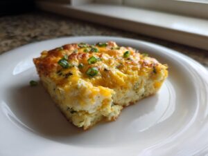 A square slice of fluffy sheet pan eggs topped with melted cheddar cheese and fresh green chives on a white plate.