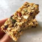 A close-up view of a chewy homemade granola bar held in hand, showing oats, almonds, and pumpkin seeds.