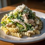 A close-up of a plate piled high with creamy chicken caesar pasta salad featuring rotini pasta, shredded chicken, and romaine lettuce.