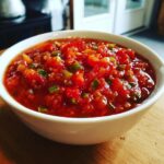 A close-up of a white bowl filled with chunky, vibrant red homemade salsa, featuring visible diced tomatoes and green peppers.