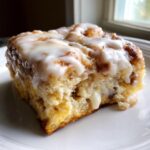 Close-up of a moist square slice of cinnamon roll casserole topped with thick white icing.