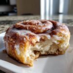 Close-up of a soft, fluffy cinnamon roll with heavy cream glaze, showing a bite taken out.