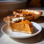 A close-up slice of classic pumpkin pie recipe topped with whipped cream on a white plate.