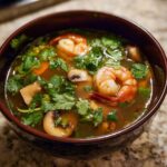 A close-up of a dark bowl filled with vibrant tom yum soup, featuring large shrimp, mushrooms, and fresh cilantro.