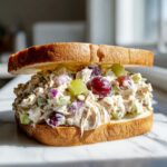 A thick, creamy chicken salad sandwich featuring grapes, celery, and red onion on toasted white bread.
