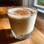 A glass of creamy, frothy coquito recipe topped generously with ground cinnamon.