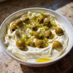 A white bowl filled with creamy dirty martini dip, topped with whole green olives and chopped pimento-stuffed olives.