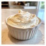 A close-up of thick, creamy garlic sauce, swirled high in a white ramekin and sprinkled with black pepper.