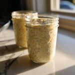Two glass jars filled with creamy overnight oats mixed with chia seeds, sitting on a marble counter in sunlight.