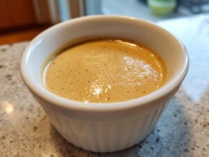 A close-up of smooth, creamy peanut sauce in a small white ramekin, showing tiny spice specks.