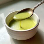 A spoonful of bright green, creamy zucchini soup drips back into a white bowl.