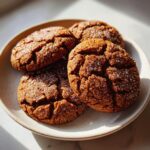 Four richly dark, crinkly ginger molasses cookies coated in sparkling sugar resting on a light ceramic plate.