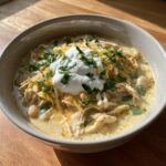 A close-up of a bowl of rich crockpot white chicken chili topped with sour cream, shredded cheese, and cilantro.