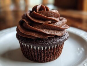 A single, moist chocolate cupcake topped with a generous swirl of dark chocolate frosting, sitting on a white plate.