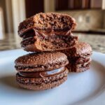 A stack of rich, dark chocolate macarons filled with glossy ganache, one cut in half showing the texture.