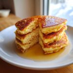 A tall stack of fluffy almond flour pancakes, cut to show the interior, drizzled generously with maple syrup.