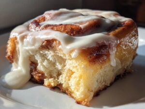 A close-up, sunlit photo of a single, fluffy cinnamon roll slice generously topped with dripping white icing.
