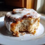A close-up of a soft, fluffy cinnamon roll with a bite taken out, generously topped with white cream cheese icing.