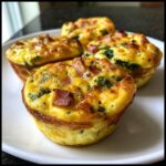 Four fluffy muffin tin egg bites topped with visible chunks of ham and spinach, served on a white plate.