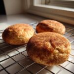 Three freshly baked Snickerdoodle cookies coated in cinnamon sugar resting on a wire cooling rack near a window.