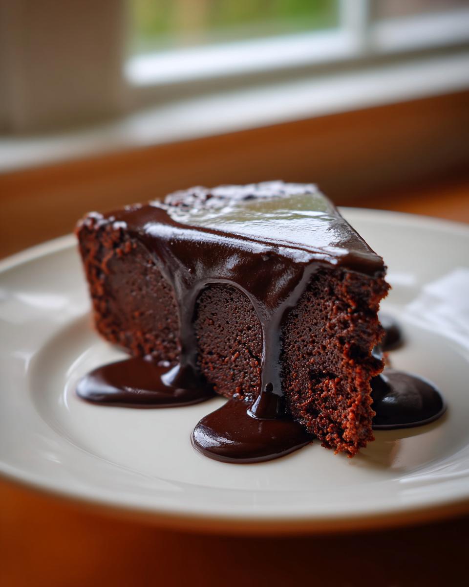 A close-up of a rich slice of flourless chocolate cake topped with glossy, dripping chocolate ganache.