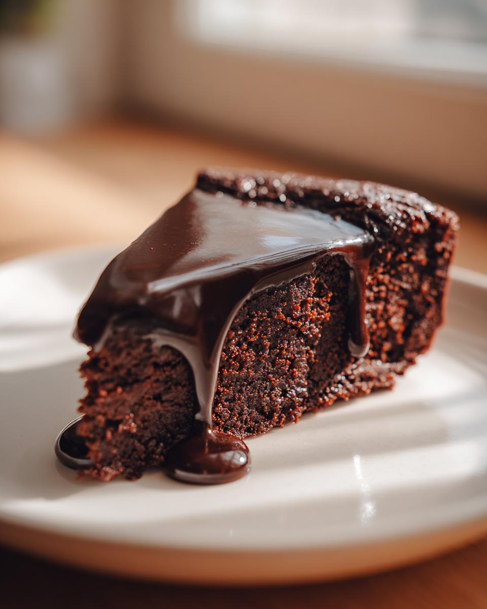 Close-up of a rich slice of flourless chocolate cake topped with glossy, dripping chocolate ganache.