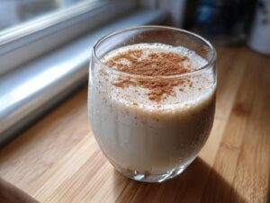 A glass of creamy, chilled coquito recipe topped generously with ground cinnamon, sitting on a wooden surface.