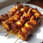 Close-up of several glazed chicken skewers resting on a white plate, showing caramelized edges and sesame seeds.