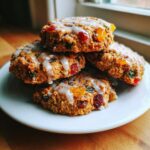 A stack of four homemade fruitcake cookies, richly studded with candied fruit and drizzled with white icing.