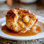 Close-up of a warm, gooey apple dumpling overflowing with spiced apples and caramel sauce on a white plate.