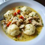 Close-up of a white bowl filled with rich chicken and dumpling soup, featuring shredded chicken, carrots, and fluffy dumplings.