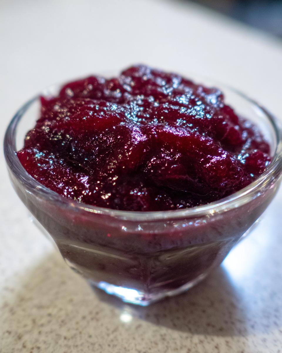 A close-up shot of thick, glistening, deep red plum jam served in a small, clear glass bowl.