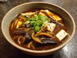 A close-up of a rich, dark broth hot and sour soup recipe, featuring tofu, shiitake mushrooms, and green onions.