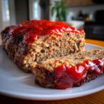 Close-up of a juicy meatloaf, sliced and topped generously with bright red ketchup glaze.