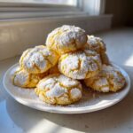 A mound of bright yellow lemon crinkle cookies generously dusted with powdered sugar, sitting on a white plate.