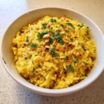 A close-up of bright yellow mediterranean rice topped with toasted pine nuts and chopped parsley in a white bowl.