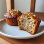 Close-up of a banana nut muffin broken in half showing the moist crumb texture and topped with chopped nuts.
